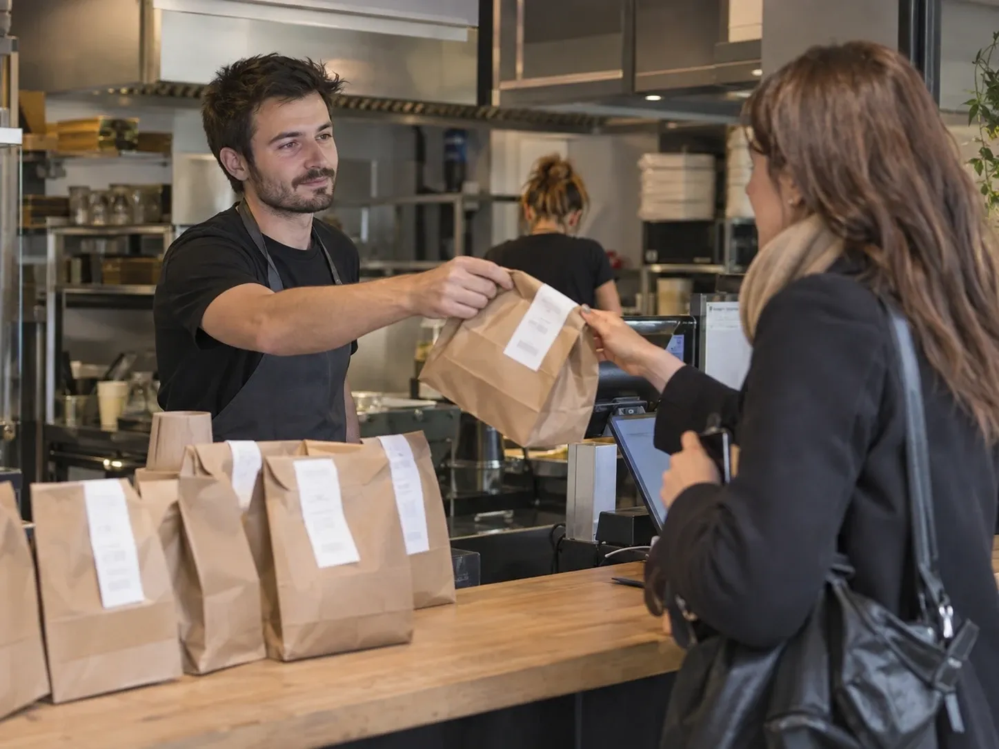 Un employé tend un sac de commande à emporter à une cliente, devant une rangée de sacs préparés.
